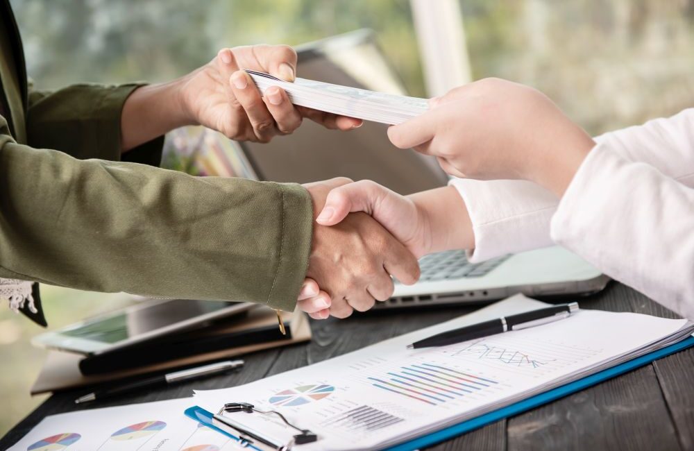 Two people shaking hands while exchanging a stack of cash, with business documents and a laptop on the table—illustrating a transparent transaction with a legal money lender in Singapore.