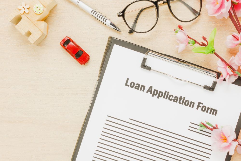 A person sits at a table looking stressed while holding a document, with a jar of coins and a small wooden house model in the foreground—highlighting the struggles of seeking a personal loan for low income Singapore residents.