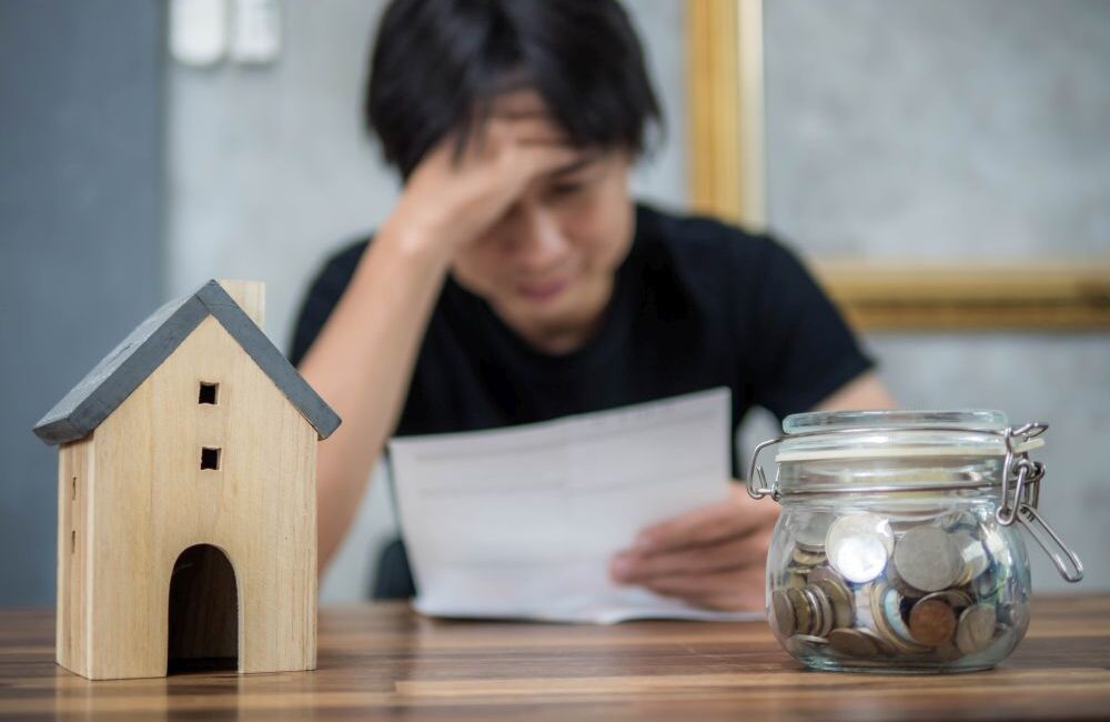 A person sits at a table looking stressed while holding a document, with a jar of coins and a small wooden house model in the foreground—highlighting the struggles of seeking a personal loan for low income Singapore residents.