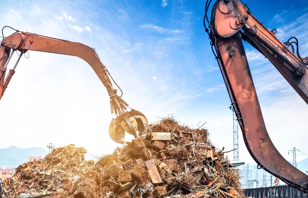Two excavators with claw attachments move a large pile of metal scrap under a blue sky at a recycling or construction site, demonstrating efficient operations that can help you understand how to choose a scrapyard in Singapore for your own recycling needs.
