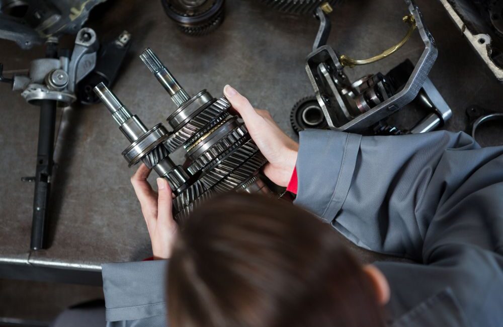 Person wearing a gray uniform examines a metal gearbox mechanism on a workbench with various gears and automotive parts nearby, demonstrating the attention to detail essential when considering how to choose a reliable gearbox specialist.