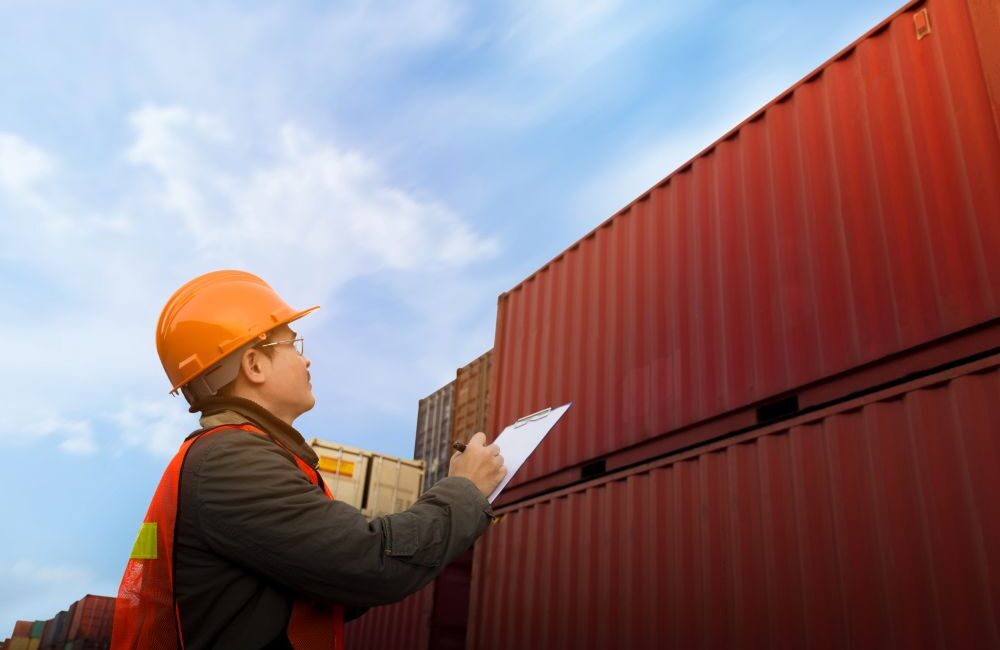 Person in safety gear holding a clipboard inspects stacked shipping containers for sale outdoors under a blue sky, highlighting what businesses should know before making a purchase.